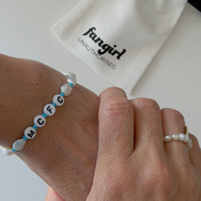 Load image into Gallery viewer, A close-up of a person's wrist wearing a beaded bracelet with white letter beads spelling 'MCFC' and light blue spacer beads. A white pouch printed with the brand name 'Fangirl Unauthorised' is visible in the background, along with another pearl ring on a finger.