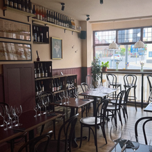 Load image into Gallery viewer, Basil&#39;s wine bar interior with tables, chairs, and wine glasses near a window.
