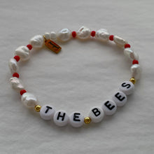 Load image into Gallery viewer, A beaded stretch bracelet featuring white baroque pearls, small red beads, gold spacer beads, and white letter beads spelling 'THE BEES'. A small gold tag is attached on the left side.
